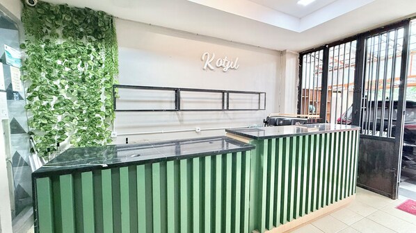A reception desk with a green and white striped design, a glass counter, and a wall with green foliage. - New Kotjil Room (Jakarta)