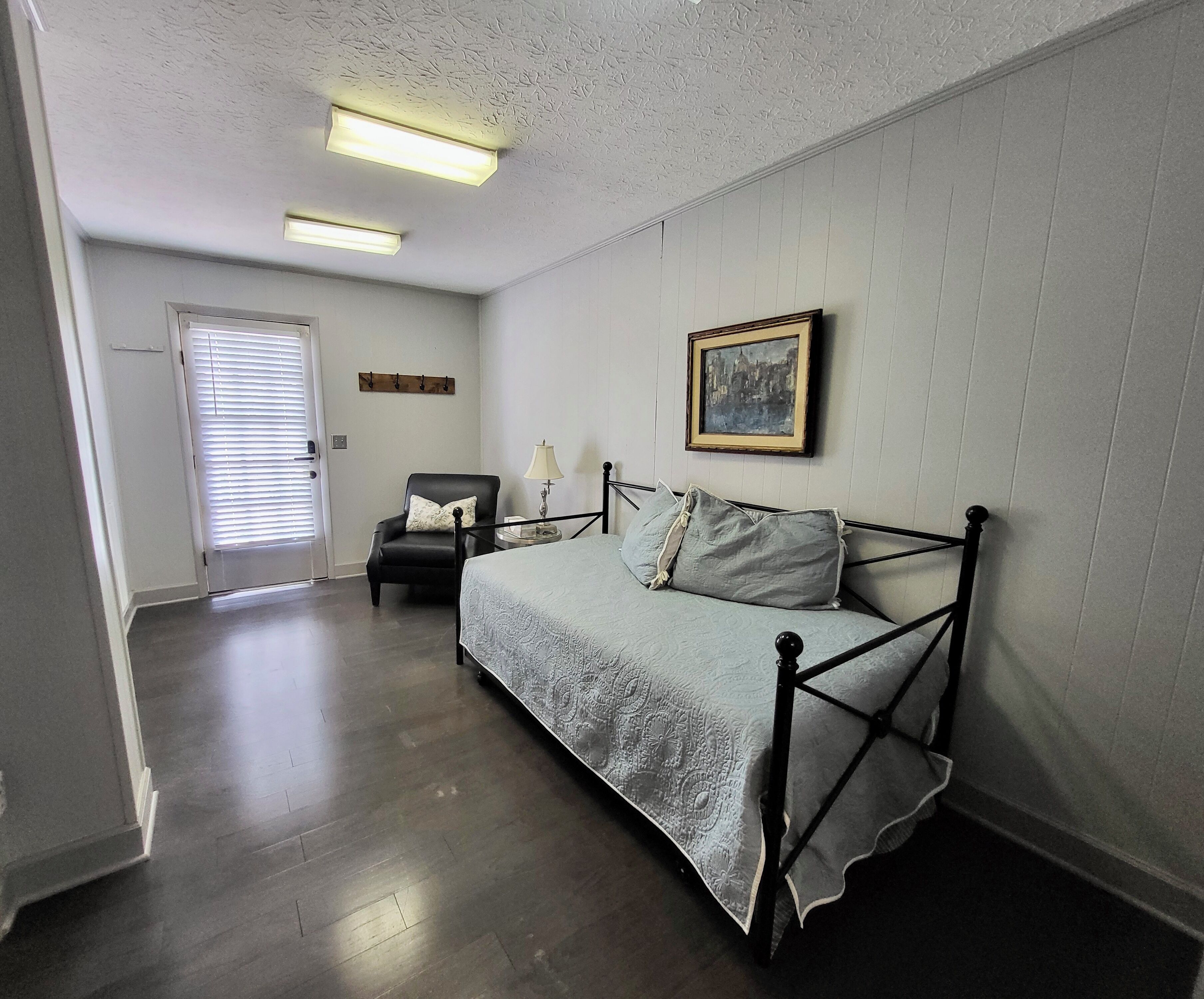 Quarto com uma cama, uma cadeira, um abajur, um quadro e uma porta.