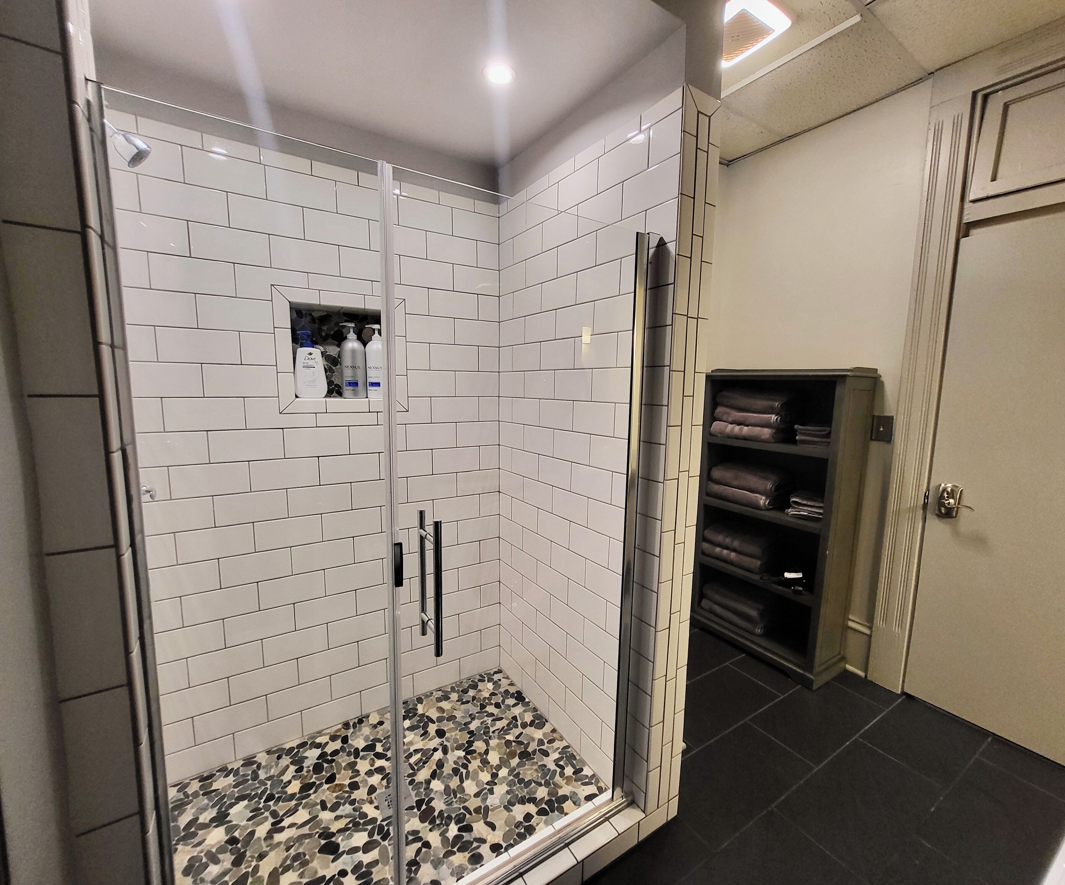 A shower with white tiles and a pebble-patterned floor, a glass door, and a shelf with toiletries.