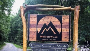 Ein Holzschild mit Berglogo und dem Text Mulberry Gap, das Annehmlichkeiten wie selbstgekochte Mahlzeiten, heiße Duschen und ein Stallgebäude auflistet.