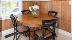 A dining room with a wooden table and four chairs, a vase with flowers, and a window with blinds.