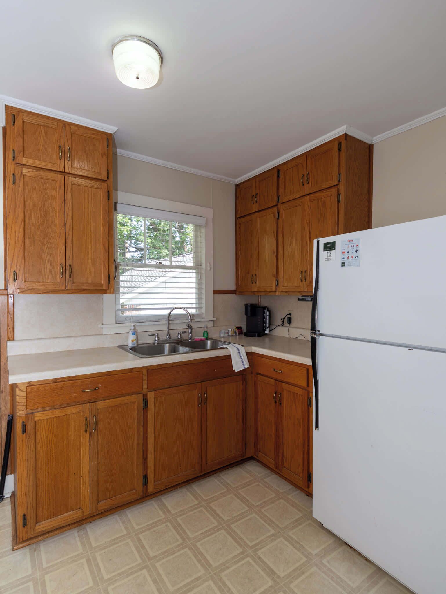 A kitchen with wooden cabinets, a white refrigerator, a window with blinds, and a tiled floor.