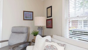 A living room with a gray armchair, a white sofa with a decorative pillow, a side table with a lamp, and framed pictures on the wall.