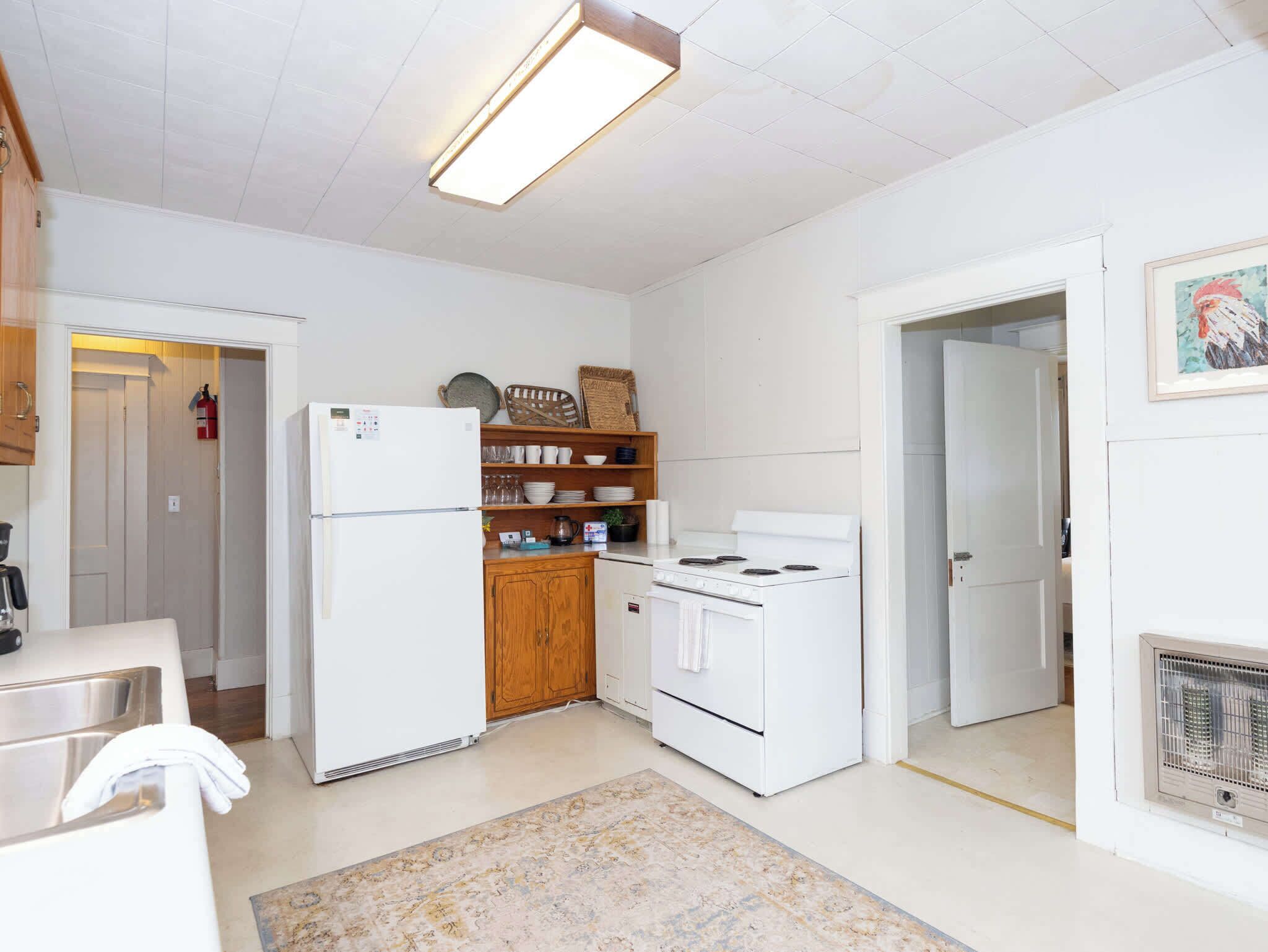 A compact kitchen with white appliances, wooden shelves, and a patterned rug.