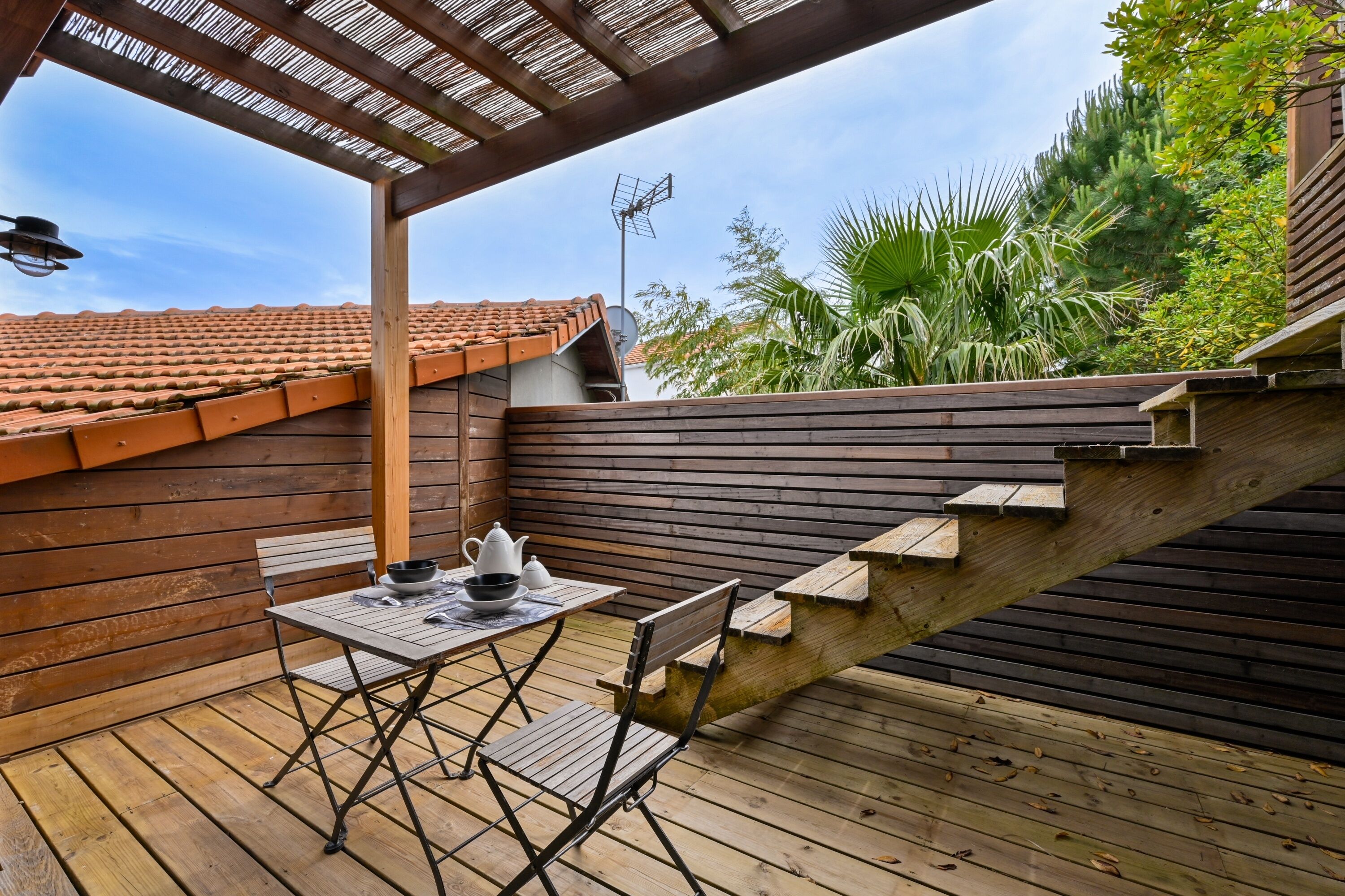 A wooden deck with a table and chairs, a pergola, and a staircase leading to a roofed area.