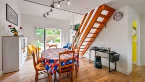 Un coin repas avec une table en bois, des chaises et une nappe colorée. Un escalier se trouve à droite, et une fenêtre offre une vue sur la verdure à l’extérieur.