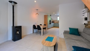 Una sala de estar moderna con chimenea de leña, un comedor con sillas, un sofá con cojines y una cocina al fondo.