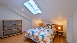 Un dormitorio con tragaluz, una cama con colcha colorida, una mesita de noche con lámpara y una estantería de madera.