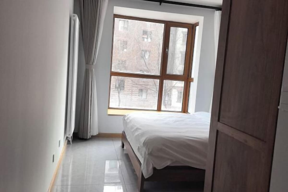 A bedroom with a large window, a bed, and a wooden wardrobe.