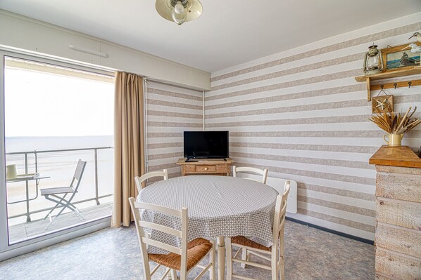 A room with a round table and chairs, a television on a wooden stand, striped wallpaper, and a view of the beach through a large window with curtains.