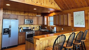 A cabin-style kitchen with wooden walls and ceiling, stainless steel appliances, and a bar area with four chairs. - Mountain View Escape - Stunning Views! Bethlehem, NH Cabin Near Skiing & Hiking (Bethlehem)