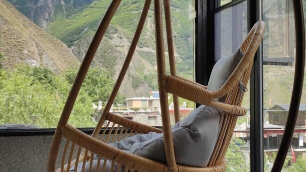Une chaise en osier avec des coussins, une fenêtre et une vue sur des collines verdoyantes.