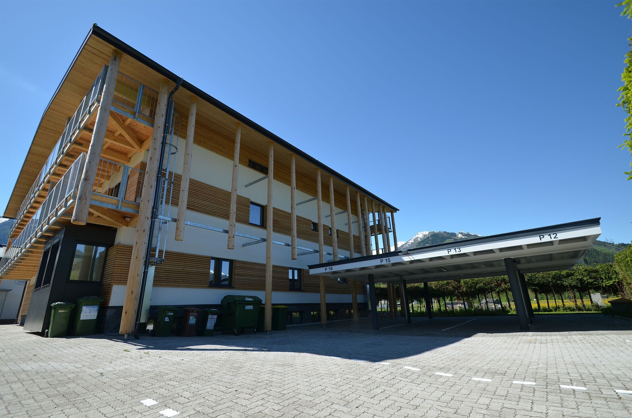 En byggnad med flera våningar, ett överbyggt entréområde, utomhusavfallsbehållare och en klar himmel.
