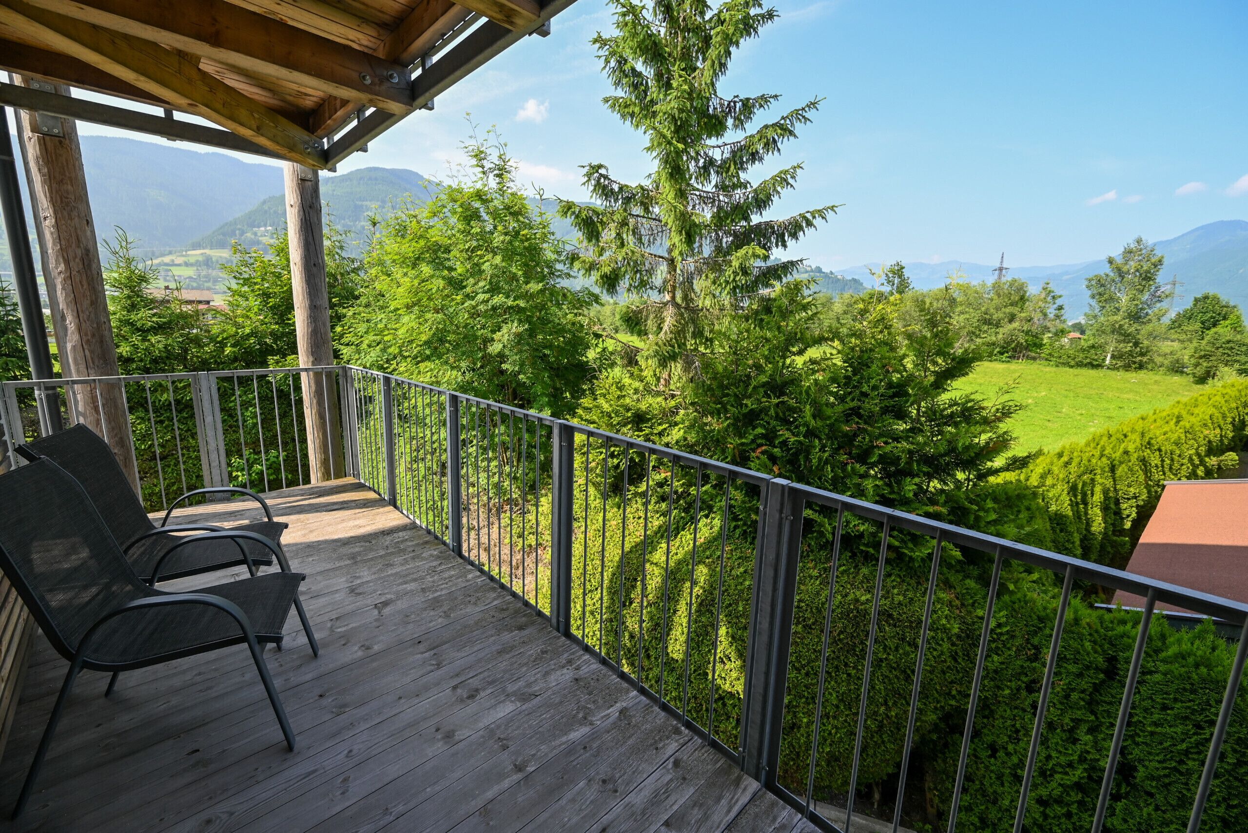 Eine Holzterrasse mit Metallgeländern, zwei Stühlen und Blick auf eine grüne Landschaft mit Bäumen und Bergen.