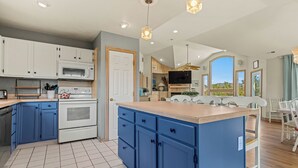 A modern kitchen with blue cabinets, a large island, a microwave, and a dining area with a view. - Spacious Pet Friendly 6-bedroom all season retreat in Corolla w/ pool and sauna (Corolla)