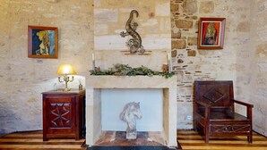 A room with a stone wall, a fireplace with a decorative animal head, a wooden chair, and two framed artworks.
