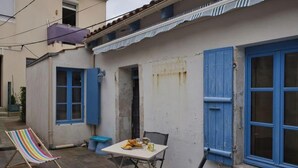 Un pequeño espacio al aire libre con una mesa y sillas, una silla de playa a rayas y una ventana con persianas azules.