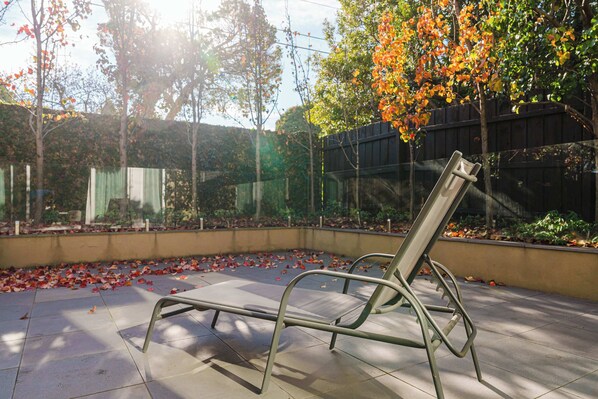 A metal bench in an outdoor area with a tiled floor, surrounded by trees and a fence.