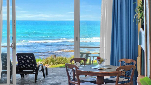 Una habitación con un gran ventanal que da a vistas del océano. En el interior, hay una mesa con sillas y un cuenco de fruta.
