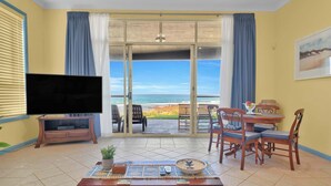 Habitación con televisión, una mesa de comedor con sillas, una mesa auxiliar y vista a la playa.