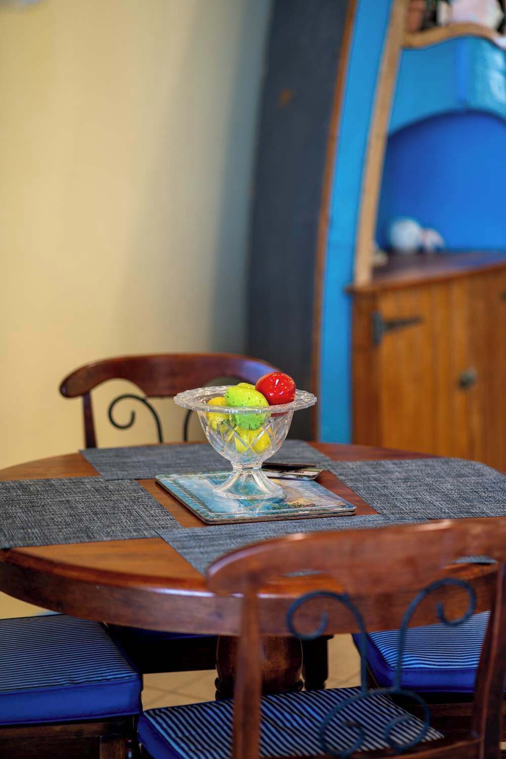 A dining table with a clear glass bowl containing fruit, a placemat, and a blue cushion on the chair.
