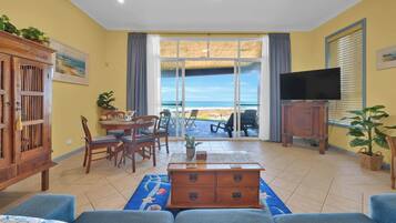 Un salon avec vue sur la plage, un coin repas et un téléviseur posé sur un meuble en bois.