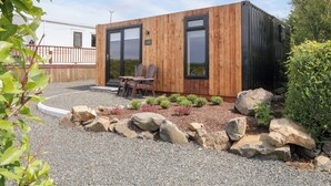 A modern wooden cabin with two large sliding glass doors, a small patio with a table and chairs, and a gravel and rock garden. - Alken The Hare (Bushmills)