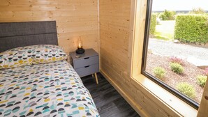 A wooden cabin room with a bed, a nightstand with a lamp, and a window with a view of a garden. - Alken The Hare (Bushmills)