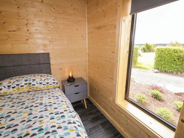 Una habitación de cabaña de madera con una cama, una mesita de noche con una lámpara y una ventana que da a un jardín.