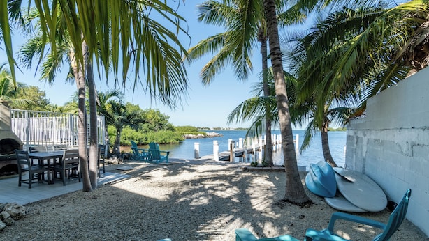 A beachside patio with a table and chairs, palm trees, and surfboards. - Sunset Key Escape Waterfront Pool Home with Dock and Sunset Views (Marathon)