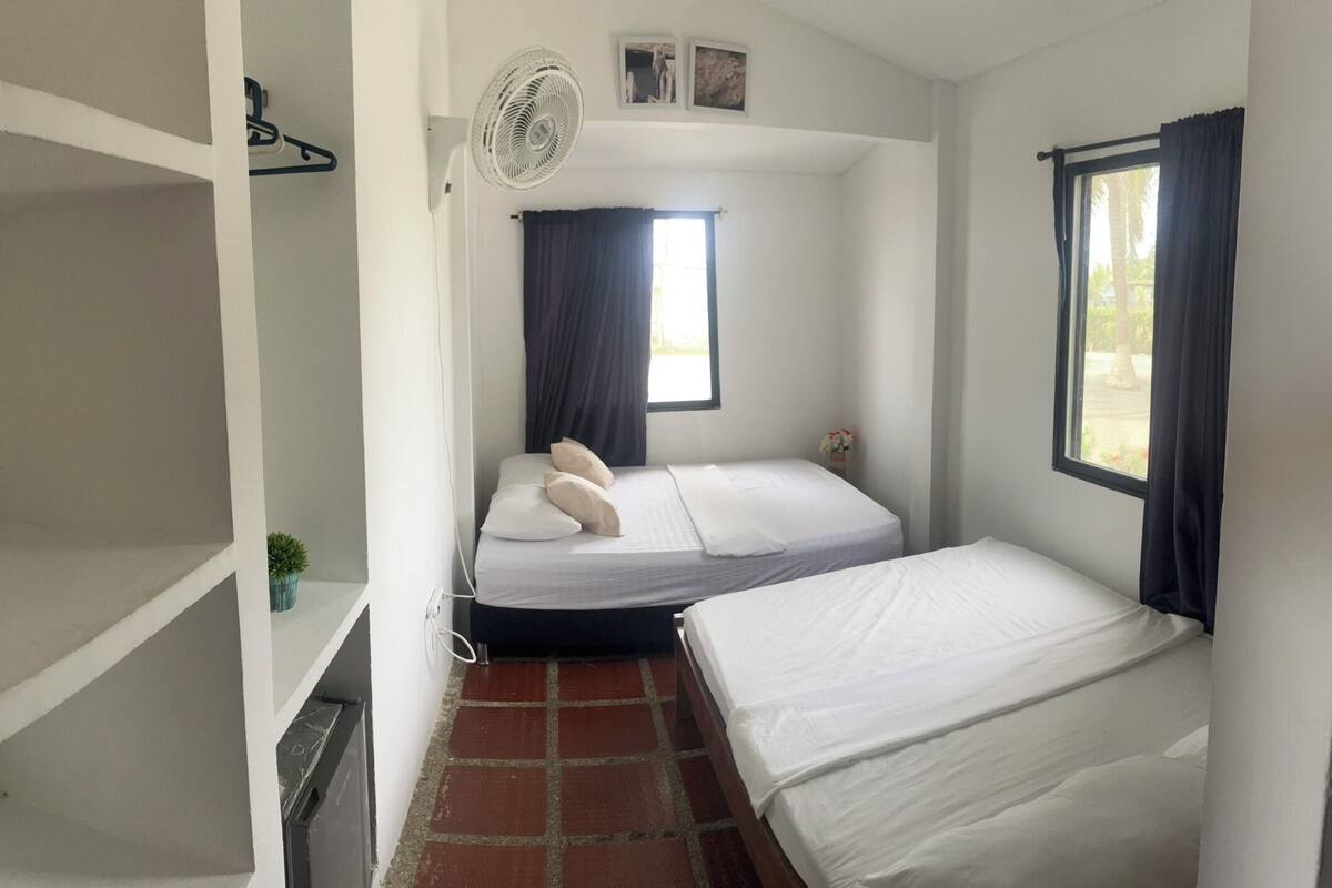 Ein kleines, sauberes Zimmer mit einem Einzelbett, einem Tischchen, einem Ventilator und einem Fenster mit Vorhängen.