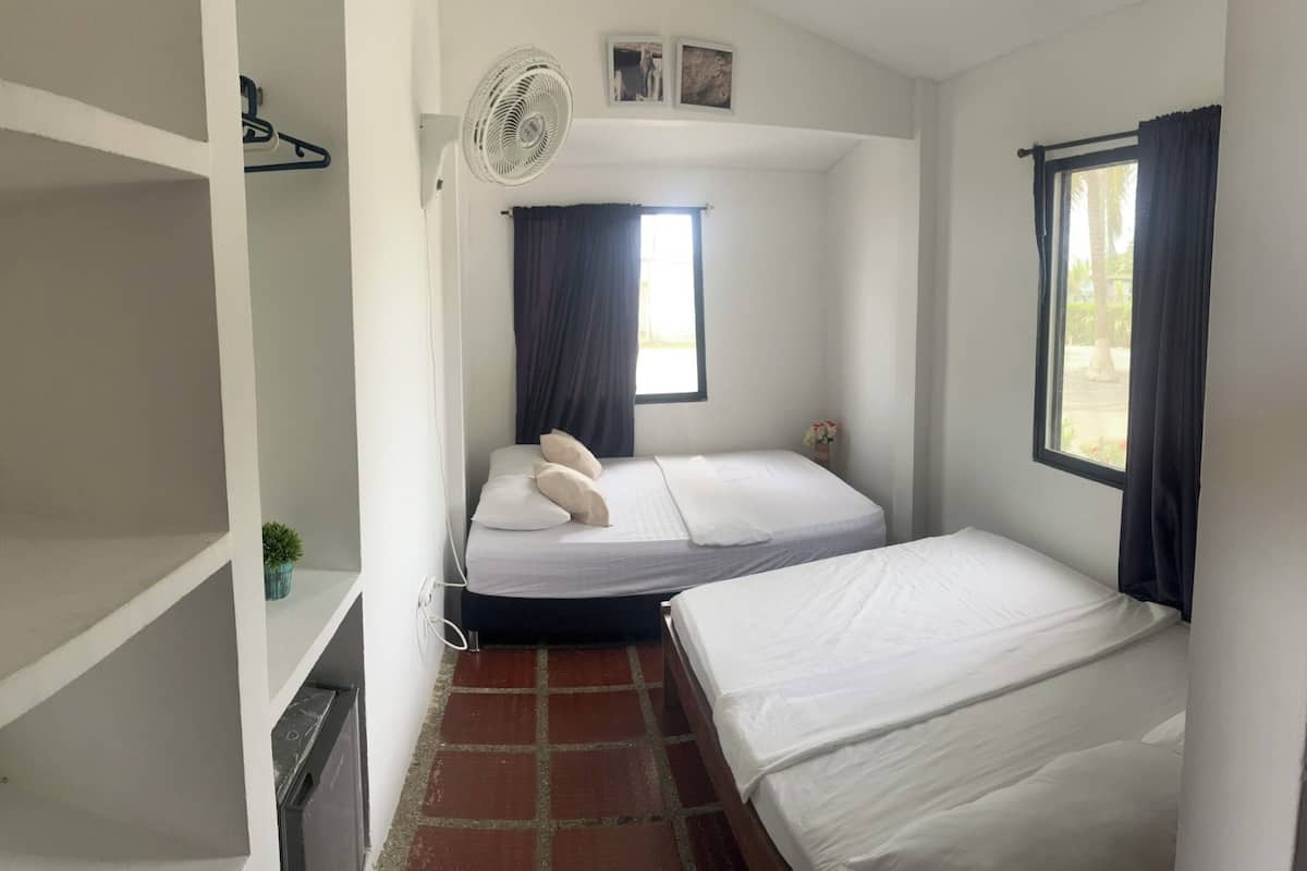Una habitación pequeña y limpia con una cama individual, una mesita, un ventilador y una ventana con cortinas.