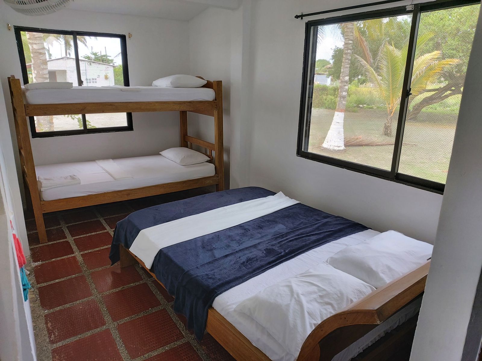 A dormitory room with bunk beds and a single bed, a window with a view of palm trees, and a tiled floor.