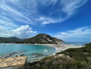 A coastal beach with clear turquoise waters, a sandy shore, and a mountainous backdrop.