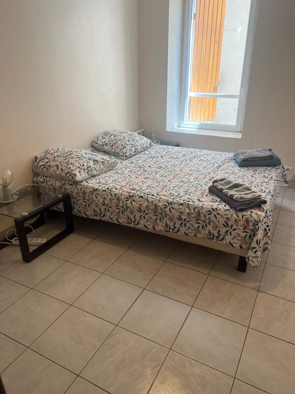 A single bed with a floral bedspread, a small bedside table, and a window with blinds.