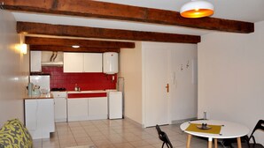 A compact kitchen with white cabinets, a red backsplash, and a wooden ceiling beam. There is a dining area with a round table and chairs.