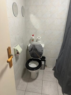 A small, tiled bathroom with a toilet, a hand dryer, a paper towel dispenser, and a toilet paper roll.