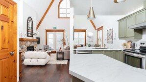 A spacious living area with a stone fireplace, a dining area with a large island, and a kitchen with green cabinets and a stainless steel range. - The Fairway: Home with Ski, Golf + Mountain Views (The Blue Mountains)