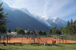 A tennis court with a mountain backdrop, surrounded by trees and benches.