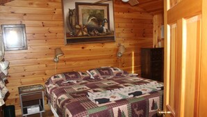 A wooden cabin bedroom with a bed, a nightstand, a lamp, a painting, and a door. - Lakeside 2-bedroom w/ AC sleeps 5. Quiet Beach in lovely Naples. (Naples)