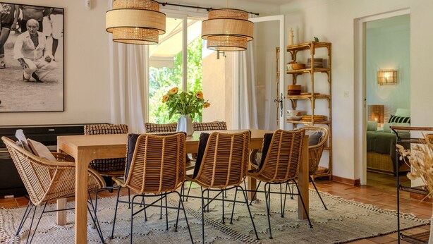 A dining area with a wooden table and wicker chairs, a vase with flowers, and a large framed photograph on the wall.