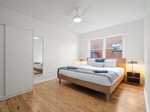 A bedroom with a wooden bed, a large window with a brick wall view, a ceiling fan, and a nightstand with a lamp.