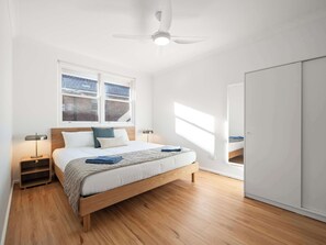A bedroom with a wooden bed, a nightstand, a ceiling fan, a window with a view of a brick building, and a wardrobe.