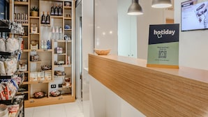 Zona de tienda y bar con estantes llenos de diversos productos, una barra con un cartel y lámparas colgantes.