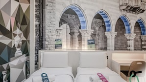 Habitación de hotel con dos camas, un diseño geométrico en la pared y vista a un edificio de piedra con arcos.