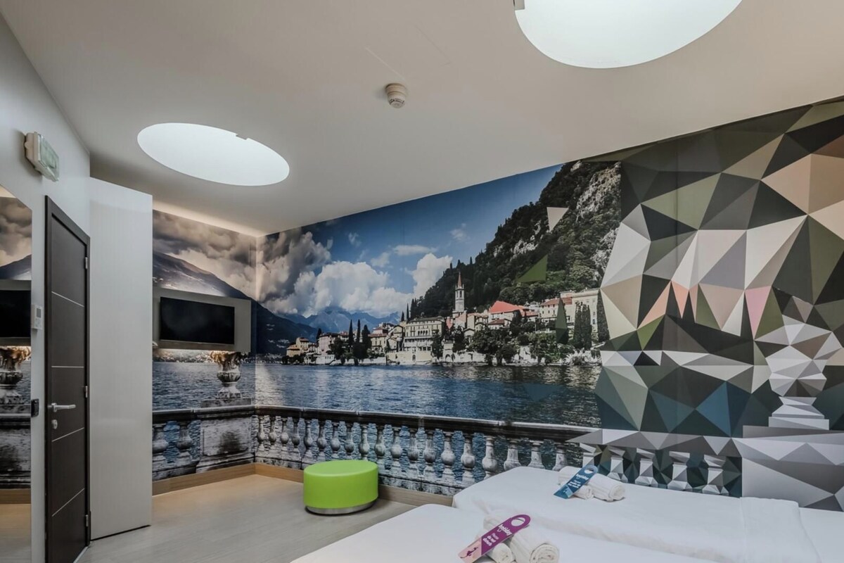 Un dormitori amb un mural gran d'una vila a la vora d'un llac, un llit, un televisor i un otomà verd.