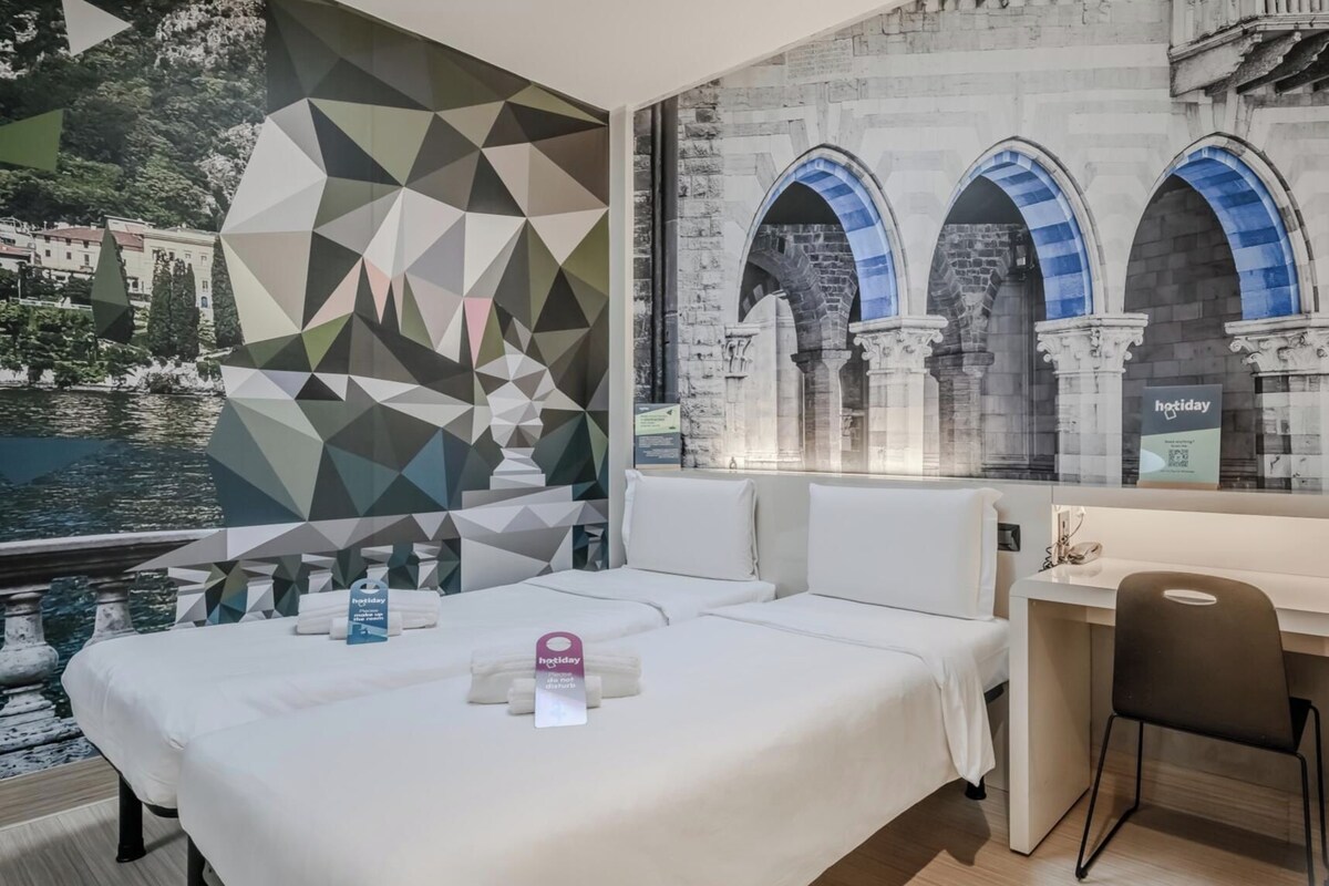 A hotel room with a bed, a desk, and a view of a historic building with arches.