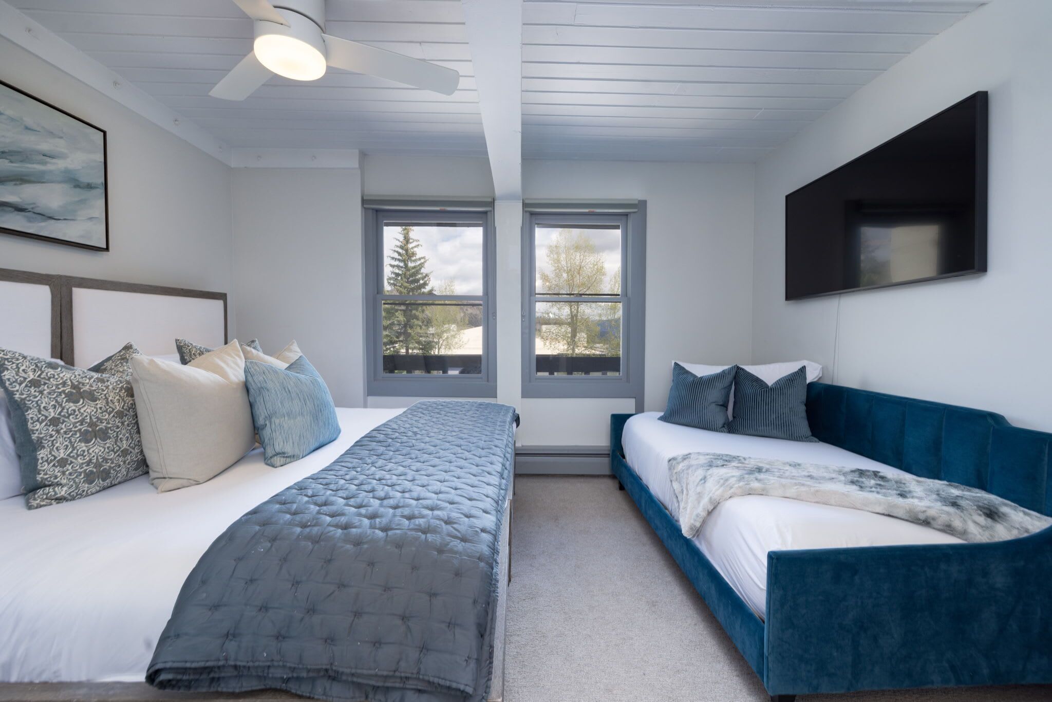 A bedroom with a bed, a sofa, two windows, a ceiling fan, and a wall-mounted TV.