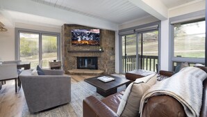 A modern living room with a stone fireplace, leather sofas, a coffee table, and large windows offering a view of the outdoors.