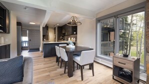 A modern kitchen with a dining area, wooden flooring, and a large window with a view of trees.